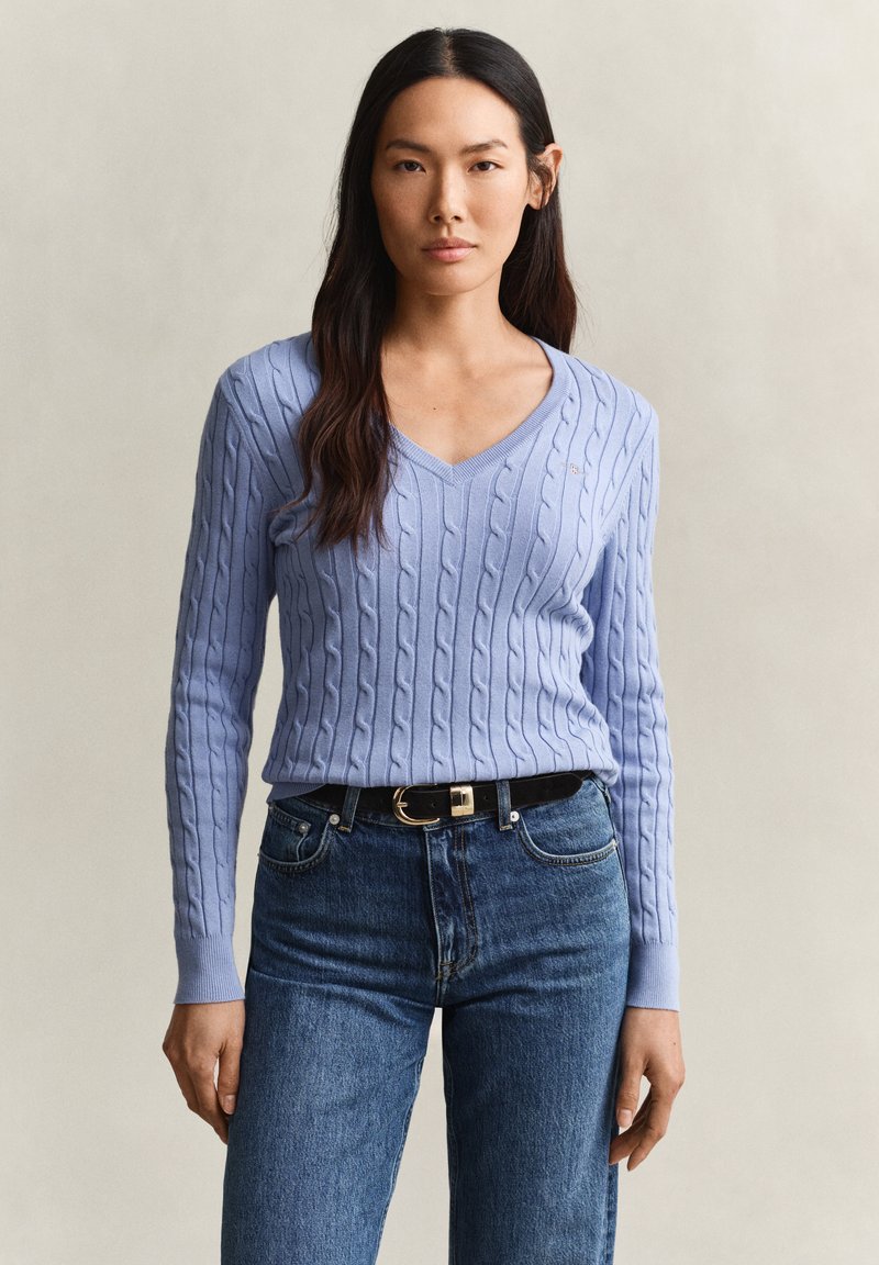 Femme aux longs cheveux foncés portant un pull bleu clair en maille torsadée à col en V et un jean bleu avec une ceinture noire, debout devant un fond uni.
