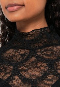 Must pitsist kaelusega must pitsist top, millel on keerukad lillemustrid ja tekstuuriga detailid, mis näitavad läbi paistvat kujundust ja pehmet viimistlust.