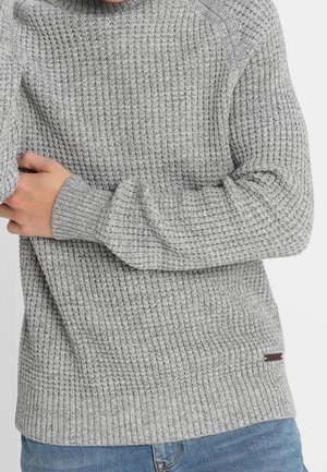 Grauer, gerippter Strickpullover mit hochgeschlossenem Kragen und langen Ärmeln, ausgestattet mit einem kleinen braunen Markenetikett an der Unterseite.