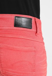 Shorts de mezclilla color coral con una etiqueta de cuero negro en la cintura, acentuados por costuras visibles y un pequeño detalle de bolsillo trasero.
