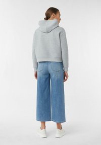 Frau mit braunem Haar im Dutt, die einen hellgrauen Hoodie, weit geschnittene, cropped blaue Jeans und weiße Turnschuhe trägt, steht vor einem schlichten weißen Hintergrund.