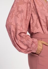 Blouse rose avec un motif floral texturé et des manches transparentes. Le tissu est léger, rehaussé d'une ceinture rose ajustée.