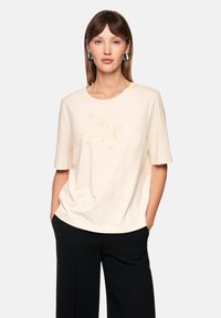 Short-sleeve cream t-shirt in soft fabric featuring a subtle gold embroidered design. Paired with wide black pants, creating a relaxed look.
