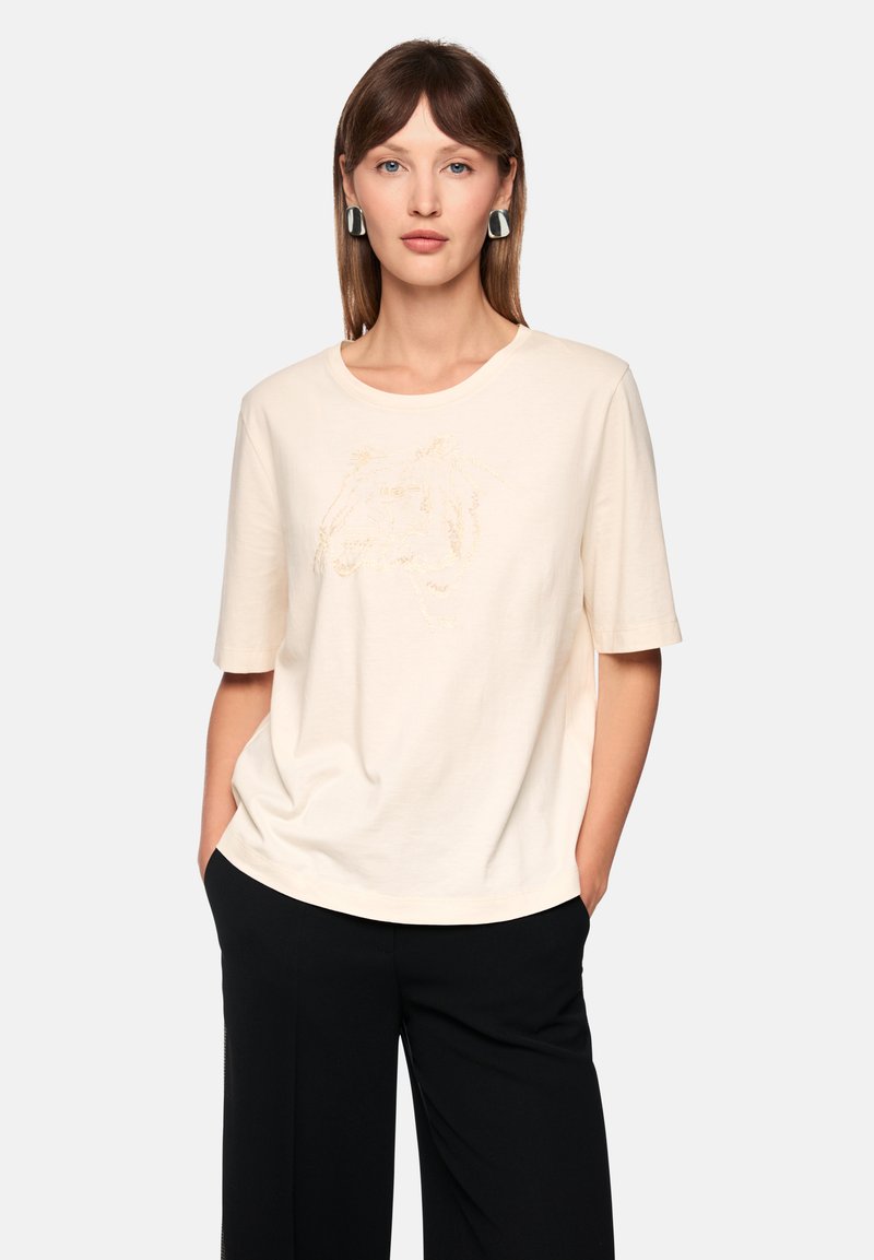 Short-sleeve cream t-shirt in soft fabric featuring a subtle gold embroidered design. Paired with wide black pants, creating a relaxed look.