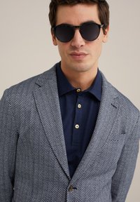 Geverifieerde blazer in marineblauw met een wit herringbone patroon, gecombineerd met een marineblauw poloshirt met drie knopen en ronde zwarte zonnebril.