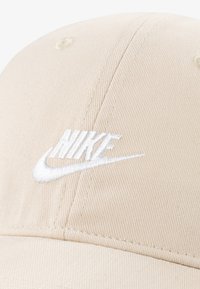 Μπεζ καπέλο μπέιζμπολ από υφασμάτινη υφή με λευκό κεντημένο λογότυπο Nike στο μπροστινό μέρος και ορατές λεπτομέρειες ραφής.