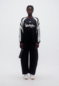 Γυναίκα στέκεται με το σώμα προς τα εμπρός φοράει μαύρο φούτερ και παντελόνι Adidas, κρατώντας μαύρο παγούρι νερού, με άσπρα αθλητικά παπούτσια σε λευκό φόντο.