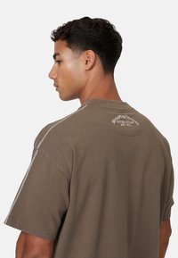 Tan oversized t-shirt met korte mouwen, een ronde halslijn en een contrasterende stiksel.details op de schouders. Achterkant ontwerp bevat tekst.