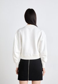 Mujer con cabello largo y oscuro que lleva puesto un sudadera blanca de manga larga y una falda negra con cremallera central sobre un fondo liso.