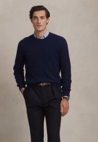 Homme portant un pull en maille côtelé bleu marine, une chemise à col rayée, un pantalon sombre, une ceinture marron et une montre, debout contre un fond beige uni.