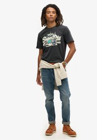 Czarny t-shirt z krótkim rękawem z grafiką "Super Dry", jasnoniebieskie dżinsy, beżowy sweter związany w talii, brązowe buty na sznurówki.