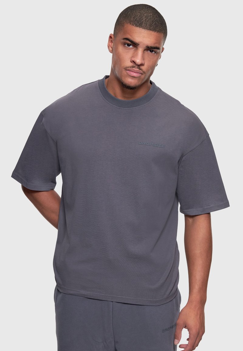 Homme portant un t-shirt gris uni oversized et un pantalon assorti, debout devant un fond gris clair avec une expression neutre.