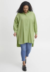Camicia oversized verde chiaro, con bottoni e maniche lunghe, con spacchi laterali, indossata sopra jeans blu aderenti e mocassini neri.