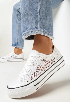 Pieds portant des baskets plateformes en dentelle crochet blanche, associées à un jean bleu clair raccourci avec ourlet effiloché, sur fond blanc.