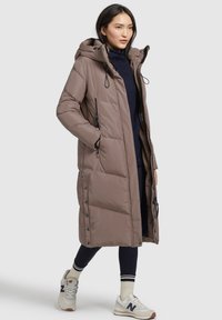 Cappotto lungo imbottito di colore taupe con cappuccio, zipper frontale e tasche laterali. Presenta dettagli in nero e chiusura a bottoni automatici.