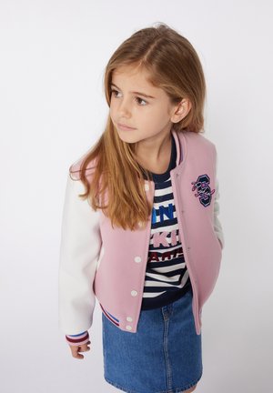 Veste de style varsity rose et blanche avec un col rond, des boutons-pression et des poignets en ribbed rayés, portée sur une chemise rayée bleu marine et blanche.
