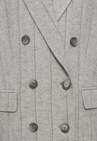 Manteau croisé gris à motif chevrons, avec six boutons noirs et deux poches à rabat.