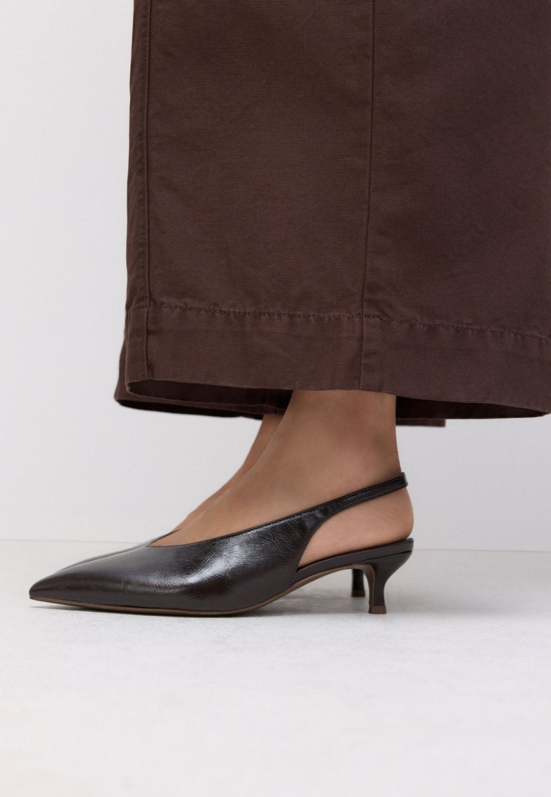 Frau trägt schwarze spitze Slingback-Kitten-Heel-Schuhe und weite braune Hosen und steht auf einem hellen Boden.