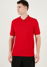 Crvena kratkarmena polo majica s ovratom na zip, izrađena od glatkog materijala, sa prilagođenim oblikom i suptilnim rebrastim detaljima na rukavima.
