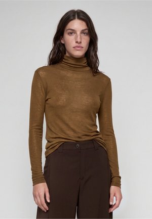 Donna con capelli lunghi e scuri che indossa un maglione marrone a collo alto e pantaloni marroni scuri, in piedi davanti a uno sfondo chiaro e semplice.