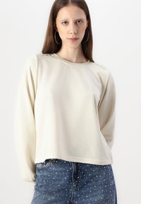 Krems krāsas garām piedurknēm krekls ar noapaļotu kakla izgriezumu, rotāts ar pērļu akcentiem plecos, savienots ar džinsu šortiem ar punktiņiem.