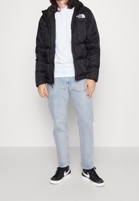 Sort puff jacket med hætte, med lynlås og logo på brystet, sammensat med lyseblå jeans og sorte sneakers.