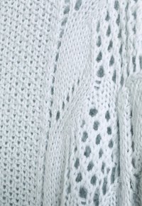 Ljusblått stickat tyg med olika mönster, inklusive en lös väv och kabelstickat mönster, vilket skapar textur och djup.