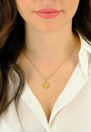 Collier pendentif en or avec motif de balance sur une chaîne délicate, porté sur une chemise blanche à col. Les cheveux encadrent doucement le visage.