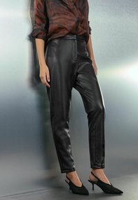 Czarne spodnie z sztucznej skóry z wysokim stanem, dopasowanym krojem i detalem szwu z przodu, w połączeniu z czarnymi butami na obcasie typu slingback na teksturowanym szarym tle.