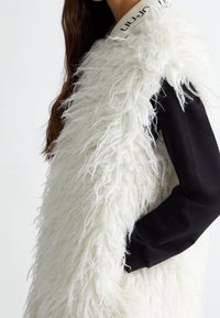 Weißer, plüschiger Weste aus flauschigem Material, darunter ein schwarzes Langarmshirt und ein strukturierter Kragen mit Beschriftung.