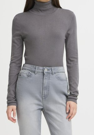 Femme portant un pull col roulé gris ajusté et un jean gris clair taille haute, debout devant un fond uni.
