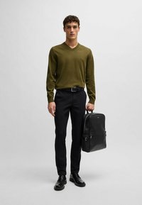 Maglione verde oliva a scollo a V, pantaloni neri su misura, scarpe di pelle nere e uno zaino in pelle nera con hardware minimale e texture liscia.