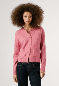 Model wearing a roze cardigan met zakken en knopen, gecombineerd met donkere jeans, met een neutrale uitdrukking tegen een effen achtergrond.