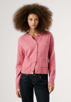 Modell trägt eine pinke Strickjacke mit Taschen und Knöpfen, kombiniert mit dunklen Jeans, und zeigt einen neutralen Gesichtsausdruck vor einem schlichten Hintergrund.