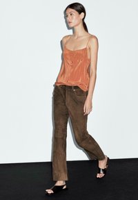 Woman walking, wearing a rust-colored satin camisole, brown suede pants, and black open-toe sandals, against a plain light background.