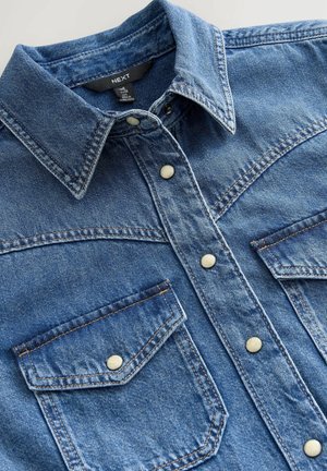 Camicia in denim con lavaggio blu, chiusura frontale a bottoni, due tasche sul petto con chiusura a scatto e cuciture a contrasto lungo le cuciture.