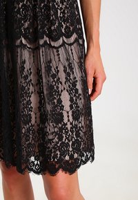 Robe en dentelle noire montrant un motif floral détaillé, un ourlet festonné et un tissu transparent. Gros plan sur une main tenant le tissu.
