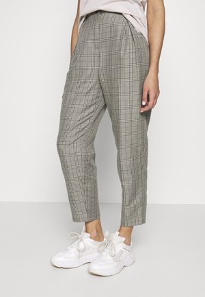 Personne portant un pantalon à carreaux gris longueur cheville et des baskets blanches, debout avec une main reposant sur le côté.