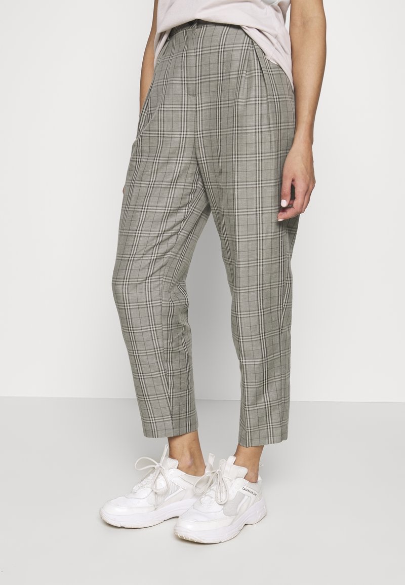 Personne portant un pantalon à carreaux gris longueur cheville et des baskets blanches, debout avec une main reposant sur le côté.
