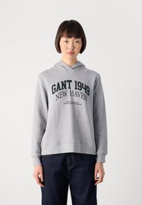 Jeune femme avec les cheveux courts et foncés portant un sweat à capuche gris avec le texte "GANT 1949 NEW HAVEN" et un jean bleu foncé, se tenant devant un fond uni.