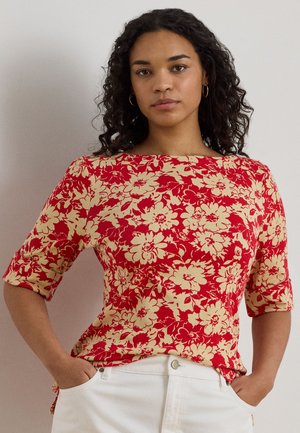 Γυναίκα με σγουρά σκούρα μαλλιά που φοράει χρυσούς κρίκους στα αυτιά και κόκκινο πουκάμισο με ανθικό σχέδιο, μαζί με λευκό παντελόνι, στέκεται με τα χέρια στις τσέπες.