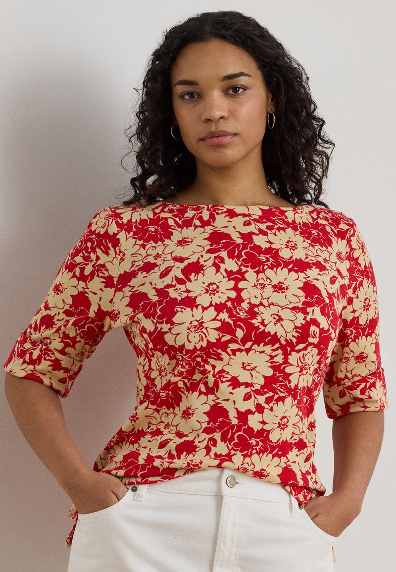 Γυναίκα με σγουρά σκούρα μαλλιά που φοράει χρυσούς κρίκους στα αυτιά και κόκκινο πουκάμισο με ανθικό σχέδιο, μαζί με λευκό παντελόνι, στέκεται με τα χέρια στις τσέπες.