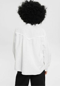 Camicia bianca oversize in lino con design a spalla e polsini abbottonati, caratterizzata da una texture morbida e una vestibilità rilassata, abbinata a pantaloni neri.