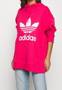 Růžová mikina s kapucí vyrobená z měkkého materiálu, která má velké bílé logo Adidas a přední kapsu, s žebrovanými manžetami a lemem.
