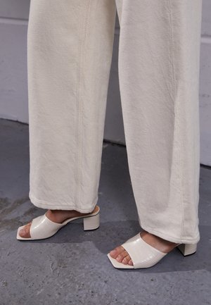 Białe mule na słupku z teksturą wytłoczonego krokodyla, zestawione z szerokimi, kremowymi spodniami z diagonalnym wzorem plecionki.