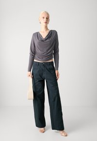 Sivi drapirani dugi rukav top kombiniran s labavim tamnozelenim hlačama. Model drži bež torbu. Glatki materijali, minimalistički dizajn, neutralne boje.