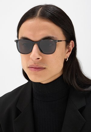 Giovane con capelli lunghi e scuri che indossa occhiali da sole neri, un dolcevita nero, un blazer nero e un piccolo orecchino a cerchio su sfondo bianco.