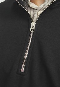 Felpa nera con zip in metallo argento e texture a coste, con un colletto a righe visibile sotto.