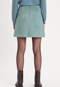 Jupe mini en suede verte avec un ourlet plat, des coutures visibles et une fermeture éclair à l'arrière. Portée avec des bottines noires et des collants opaques.