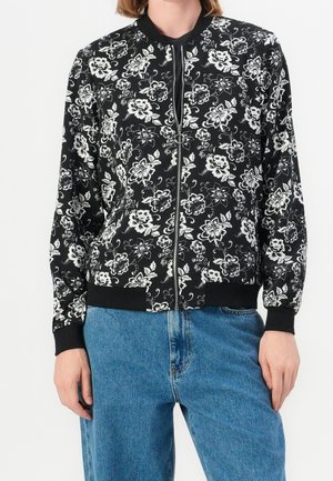 Persoon draagt een zwart jack met ritssluiting en een wit bloemenpatroon, met geribde manchetten, gecombineerd met blauwe spijkerbroek tegen een effen achtergrond.